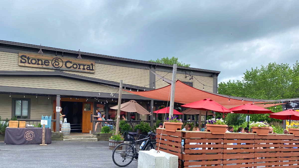 A one-story building with light brown paneling and sign that says Stone Corral in an old West-style font. In front are picnic tables with red umbrellas.