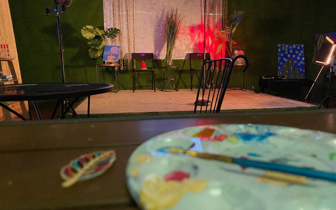 A painter's palette foregrounds a stage set with red lighting, easels, and plants.